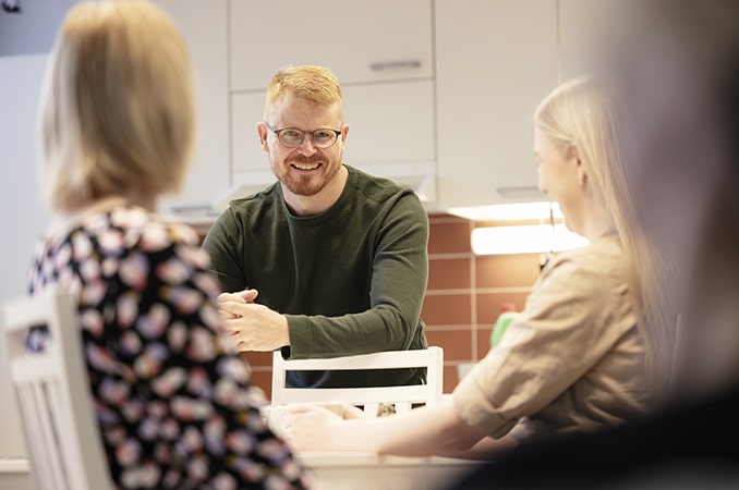 Mies istuu kahden nuoren kanssa keittiössä ja hymyilee.