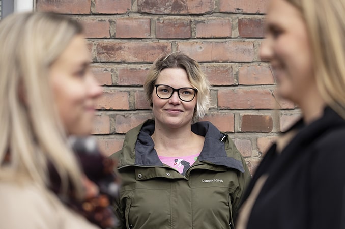 Kolme nuorta naista, joista keskimmäinen nojaa tiiliseinään. Keskimmäisen ksvot näkyvät tarkkoina, sivuilla olevien epätarkkoina.