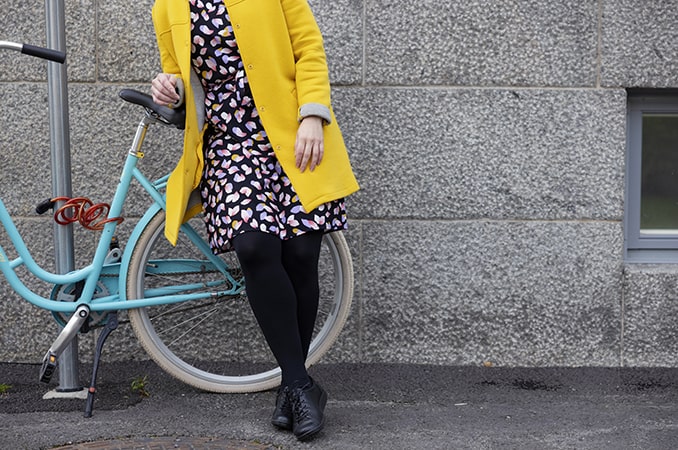 NAinen keltaisessa takissa nojaa seinään turkoosi polkupyörä takanaan.