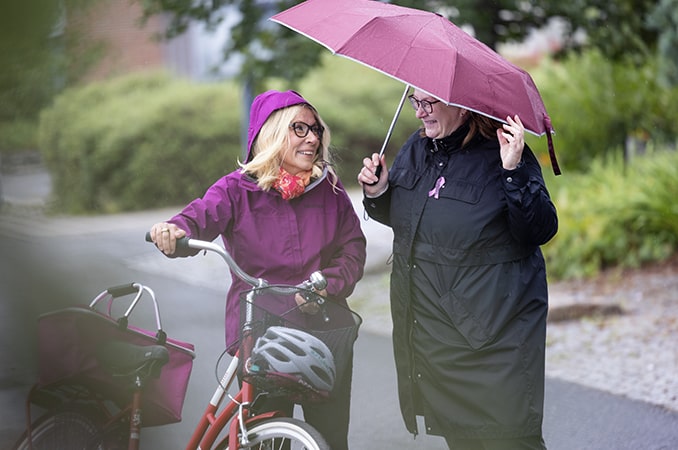 Nainen taluttaa pyörää ja hymyilee naiselle, joka on sateenvarjon alla.