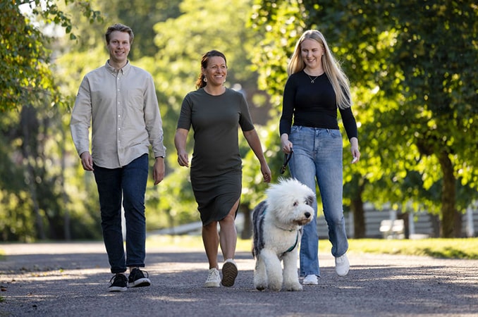 Kaksi naista ja mies kävelevät puistossa. Nainen taluttaa koiraa.