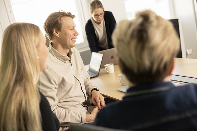 Mies ja kolme naista palaverissa. Mies hymyilee.